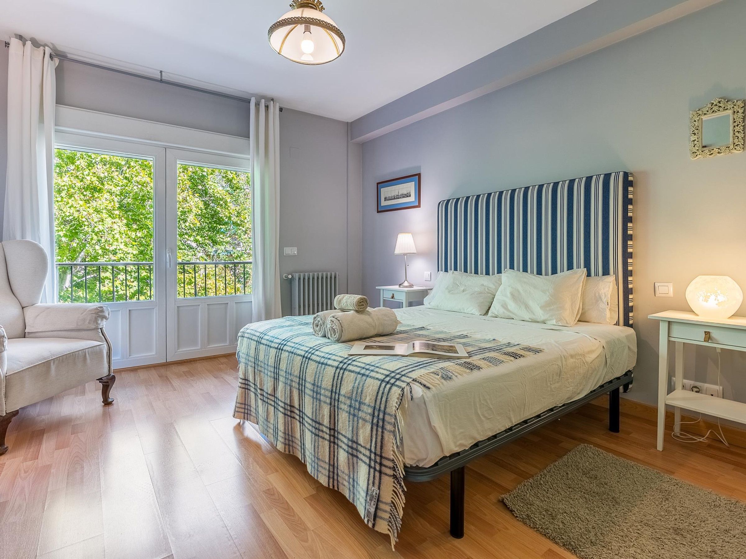 A bedroom with a bed, a chair, a bedside table, a lamp, and a view of greenery through the window.