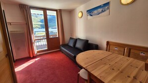 Ein Zimmer mit Holztisch, einer Couch, einem roten Teppich, einem Kühlschrank und einem fensterbepflanzten Fenster.