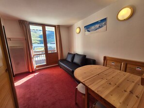 Ein Zimmer mit Holztisch, einer Couch, einem roten Teppich, einem Kühlschrank und einem fensterbepflanzten Fenster.