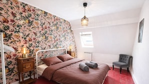 A bedroom with a floral wallpaper, a bed with brown bedding, a nightstand with a lamp, a chair, and a window. - La Maison Créative (La Madeleine)