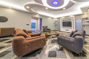 Ein modernes Wohnzimmer mit einem Ecksofa, einem grauen Sessel, einem Flachbildfernseher und einer Decke mit runden Leuchten.