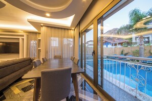 A modern living room with a dining table, sofas, and a large window overlooking a swimming pool.