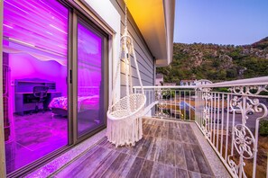 A balcony with a macrame chair, a sliding glass door leading to a room with a desk and chair, and a view of a hillside.