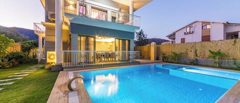 A modern two-story house with a swimming pool, a balcony with outdoor furniture, and a well-maintained garden.