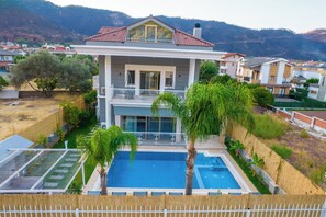 A two-story house with a swimming pool, surrounded by greenery and mountains.
