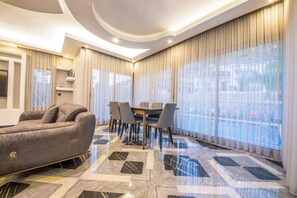 A modern living room with a sofa, a dining table with chairs, and large windows with curtains. - Daisy Villa Icmeler Marmaris Private (Marmaris)