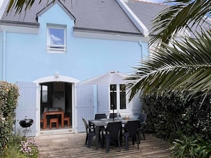 Ein hellblaues Haus mit überdachter Terrasse, Grill und Esstisch mit Stühlen.