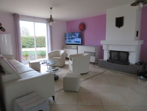 Ein Wohnzimmer mit einer Couch, Sesseln, einem Couchtisch, einem Flachbildfernseher, einem Kamin und einem Fenster mit Blick auf Grünflächen.