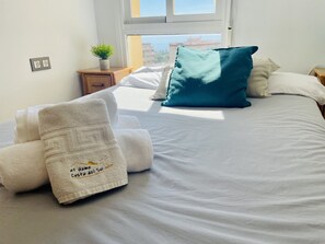 A neatly made bed with white linens and a teal pillow, a small wooden nightstand with a plant, and a window with a view of buildings. - Lighthouse 77 Apartment by At Home Costa del Sol (Torrox)