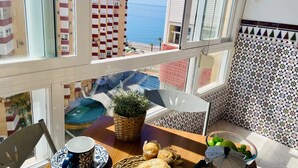 En balkon med udsigt til havet, et bord med en kop, brød, en plante og en skål med frugt.