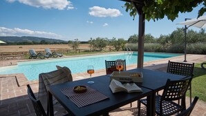 Mesa à beira da piscina com cadeiras, um livro e copos de vinho, com vista para a piscina e o campo.