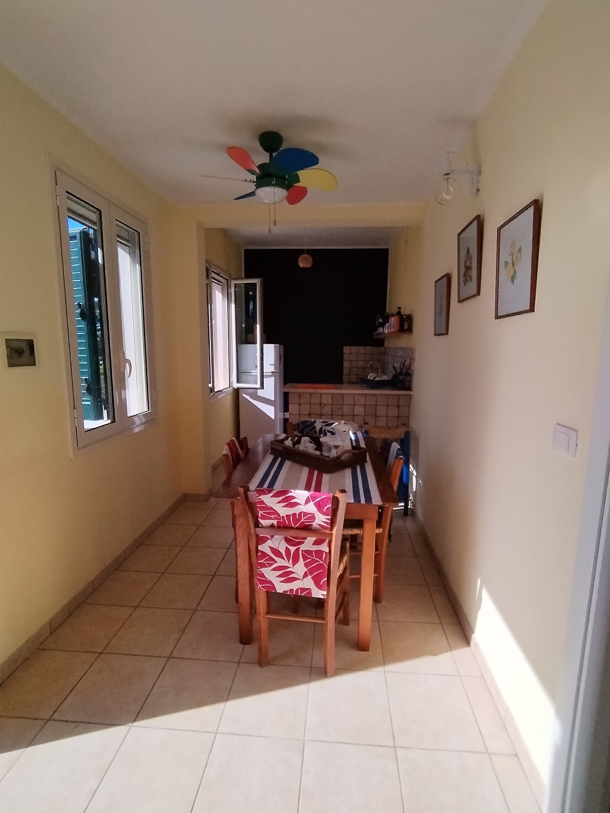 A narrow hallway leading to a dining area with a ceiling fan, a refrigerator, and a tiled floor.