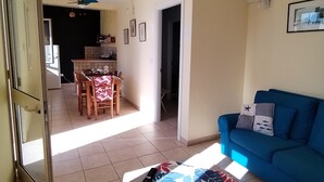 Una sala de estar con un sofá azul, una mesa de centro con mantel floreado, un ventilador de techo y una cocina al fondo.