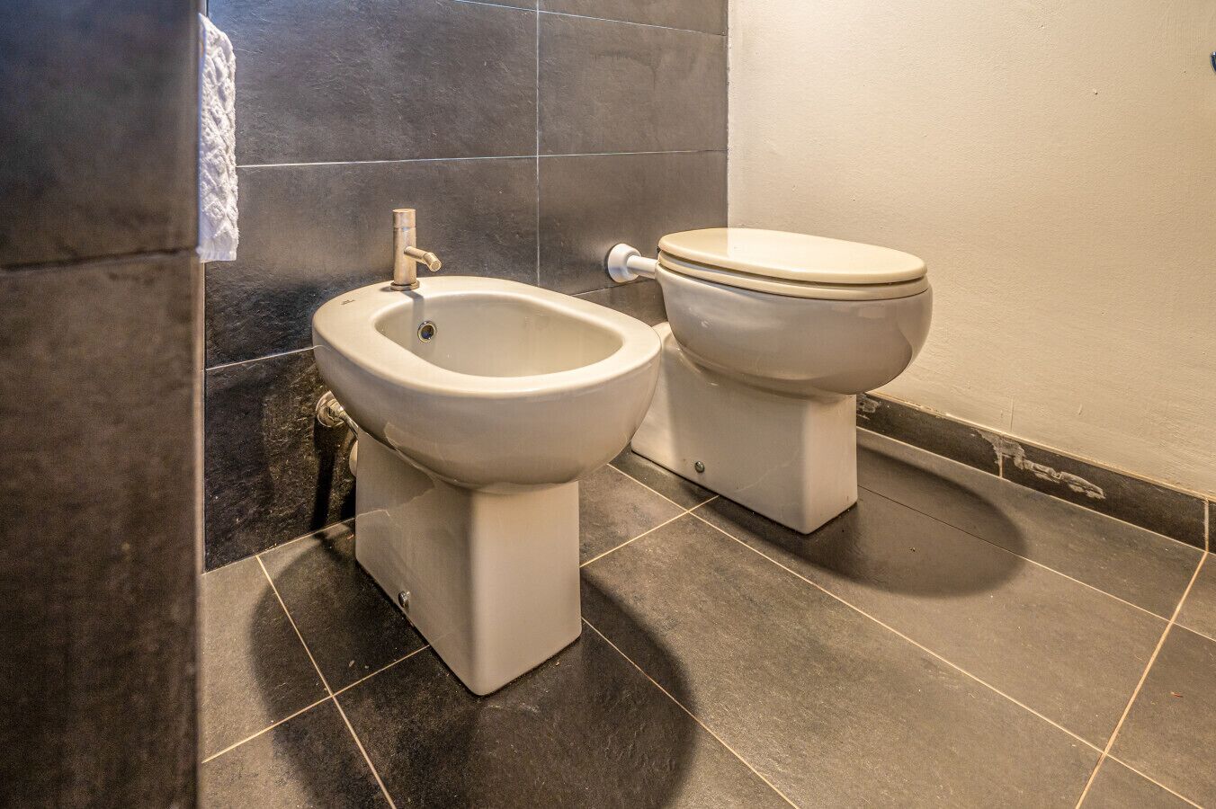A bathroom with two toilets and a bidet, tiled floor, and a towel hanging on the wall.