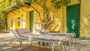 Two metal beds with pillows, a hanging lantern, and a tree in front of a yellow building with green doors.