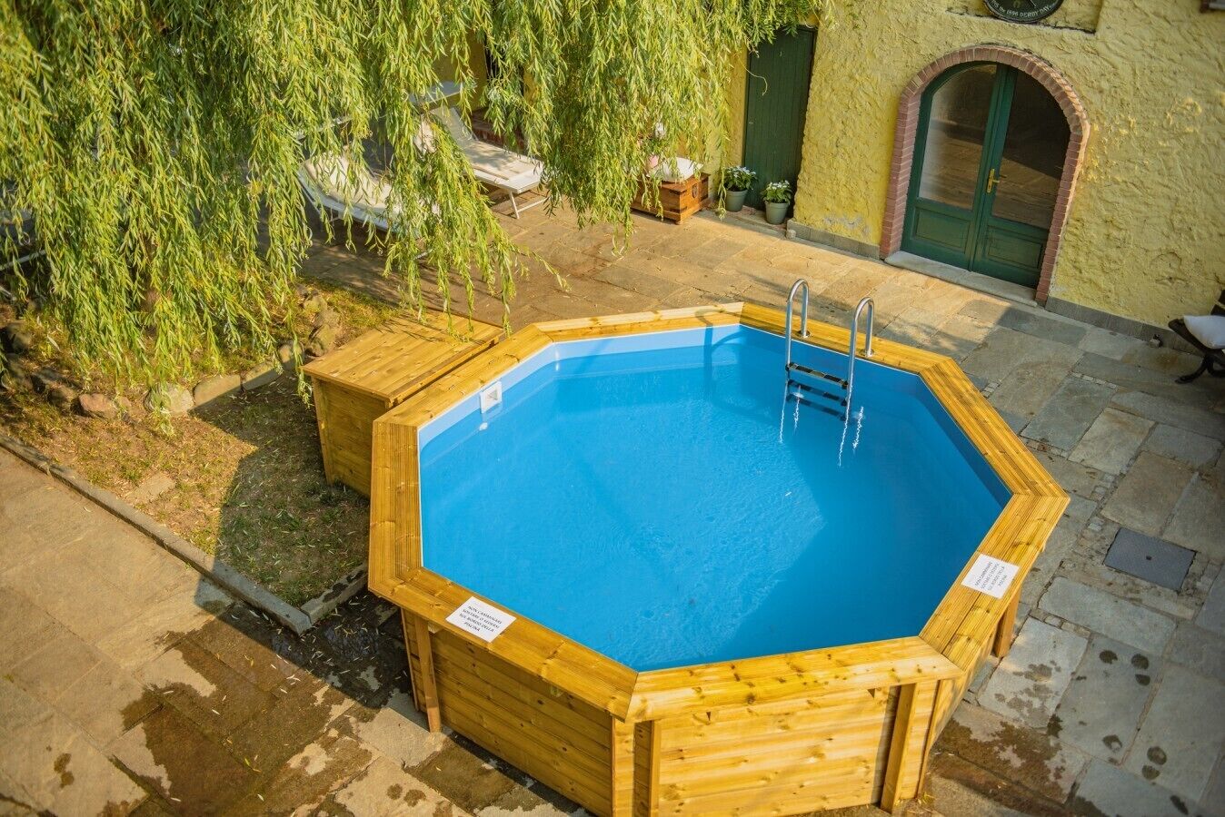 Una piscina de madera octogonal con escalera, rodeada de baldosas de piedra y una cubierta de madera.