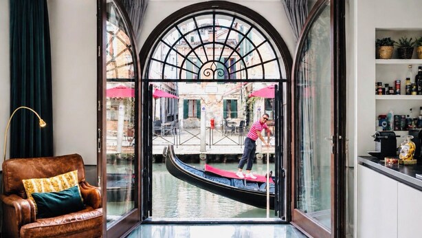 A room with a view of a canal, a gondola, a leather armchair, and a shelf with various items. - Viola Suite Luxury Canal View Central Venice (Venezia)