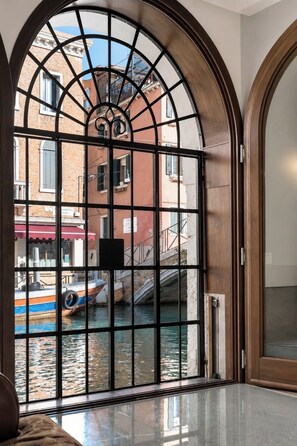 Une vue à travers une grande fenêtre en arc surmontée d’une grille métallique, donnant sur un canal où l’on aperçoit des bateaux et des bâtiments.