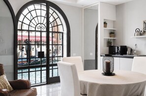 Un intérieur moderne comprenant un coin repas, une cuisine avec des meubles blancs et une grande fenêtre en arc d’où l’on peut admirer un plan d’eau.