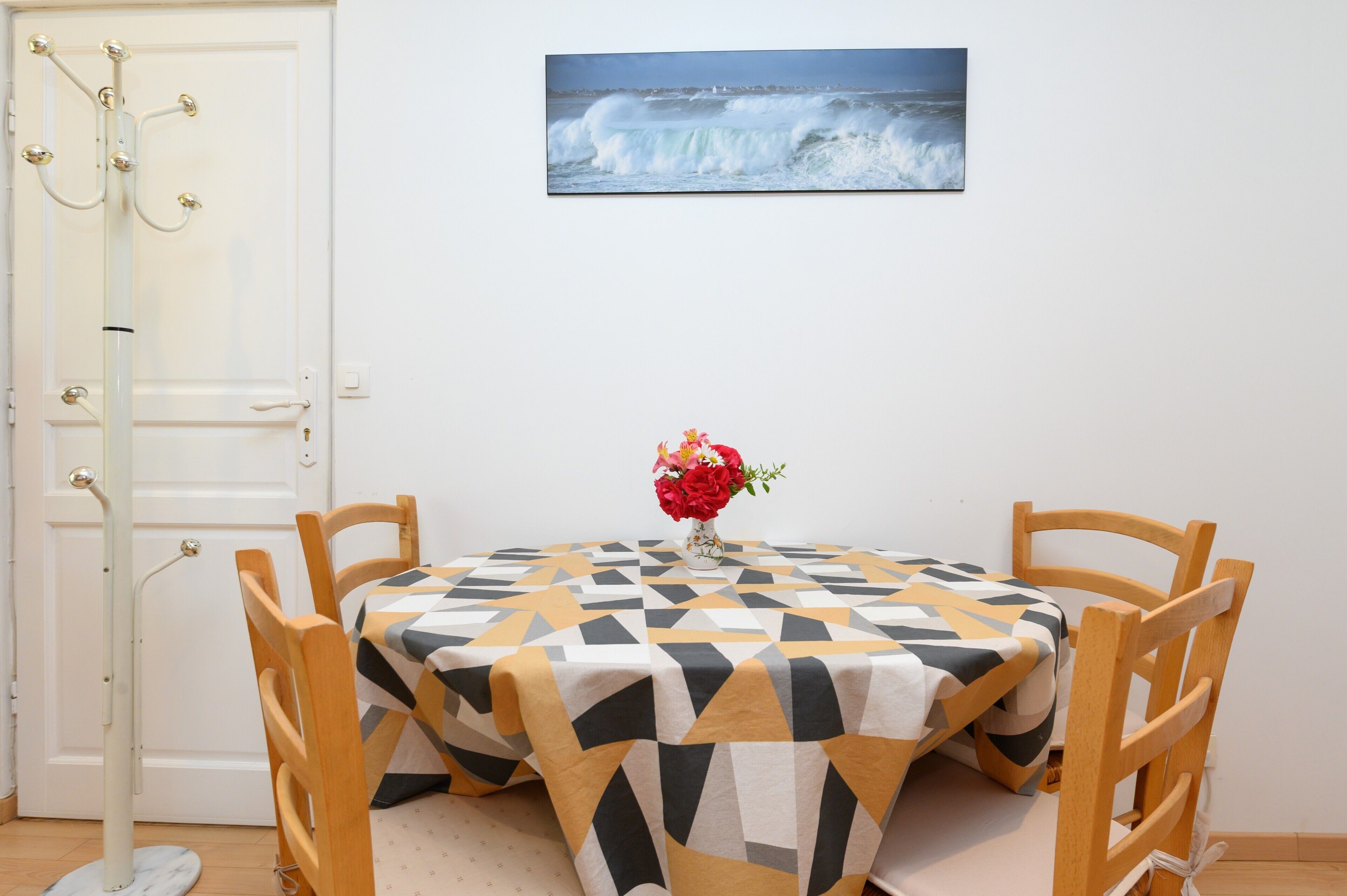 Un coin repas avec une table dressée pour deux personnes, une chaise, un porte-manteau et un tableau encadré au mur.