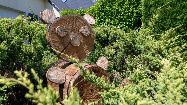 Una escultura de madera que representa un oso en un jardín con follaje denso y una casa al fondo.