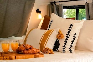 A bed with pillows, a bedside table with croissants and orange juice, and a window with curtains.