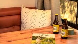 Una mesa de madera con una revista, dos botellas y una lámpara negra en una habitación con una ventana y un sofá de cuero marrón.