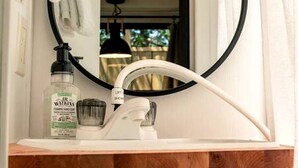 A bathroom with a white sink, a round mirror, a bottle of hand soap, and a towel hanging on the wall. - Incredible Caravan with Fire Pit and Games in Deseronto, Ontario (deseronto)