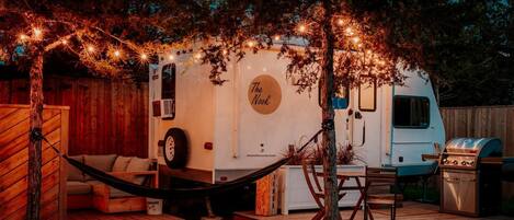 Una caravana recreativa con exterior blanco, un letrero que dice 'The Nook', una parrilla y una hamaca.
