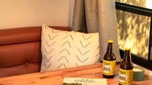 A wooden table with a magazine, two bottles, and a black lamp in a room with a window and a brown leather couch. - Incredible Caravan with Fire Pit and Games in Deseronto, Ontario (deseronto)