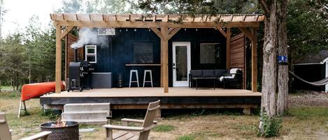 Una terraza de madera con una parrilla, asientos exteriores y un área cubierta con una puerta.