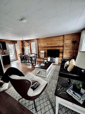 A modern living room with a wooden wall, a black leather sofa, a coffee table, and a dining area with chairs and a table. - Picturesque Cottage with Spacious Large Backyard and BBQ in Canada (Brigus South)