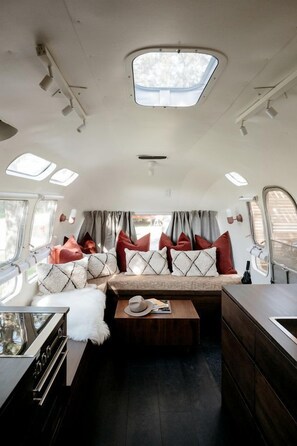 Un intérieur de camping-car compact comprenant un coin cuisine, un espace salon avec des coussins, une petite table et une fenêtre avec des rideaux.