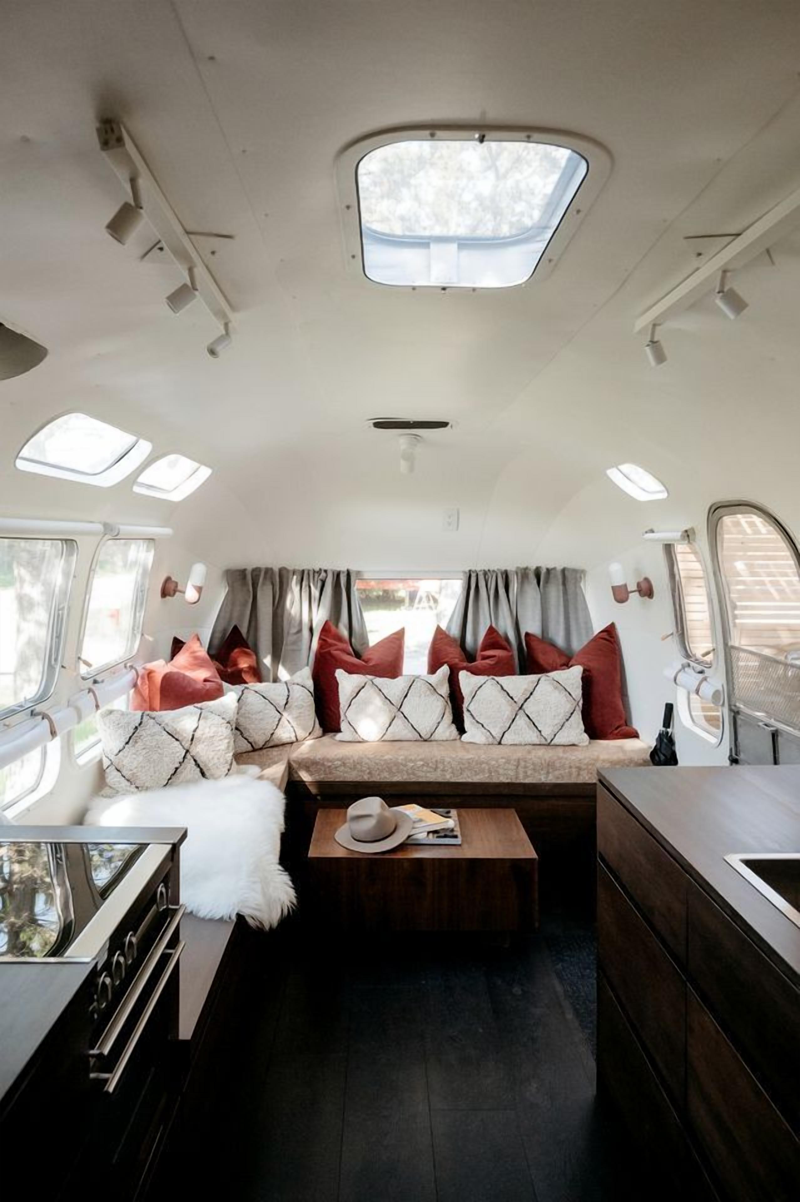 A compact RV interior with a kitchenette, a seating area with cushions, a small table, and a window with curtains.