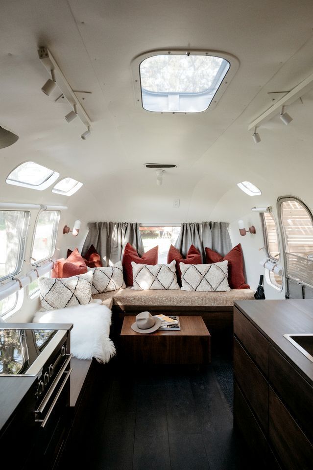A compact RV interior with a kitchenette, a seating area with cushions, a small table, and a window with curtains.
