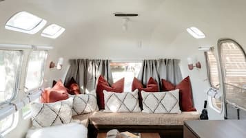A compact RV interior with a kitchenette, a seating area with cushions, a small table, and a window with curtains.