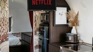 Ein kompaktes Zimmer mit Bett, Küchenzeile, einem an der Wand montierten Fernseher mit dem Hinweis 'NETFLIX' und einem gerahmten Schild mit der Aufschrift 'Wifi'.