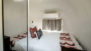 Ein kleines Zimmer mit Bett, Fenster mit Vorhang, Wandgerät zur Klimatisierung und einem kleinen Oberlicht.
