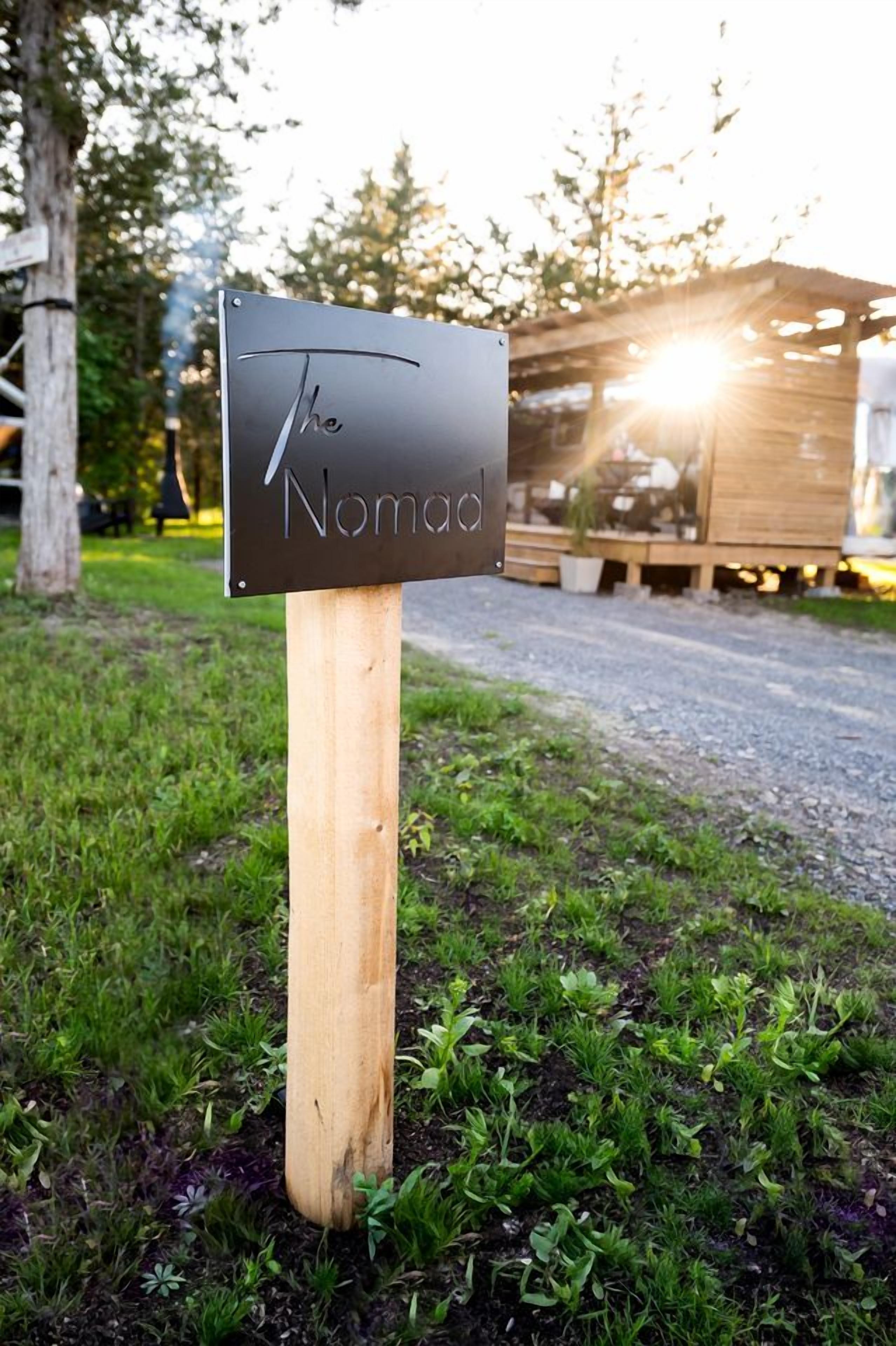 A sign with the text 'The Nomad' on a wooden post in a grassy area with trees and a wooden structure in the background.
