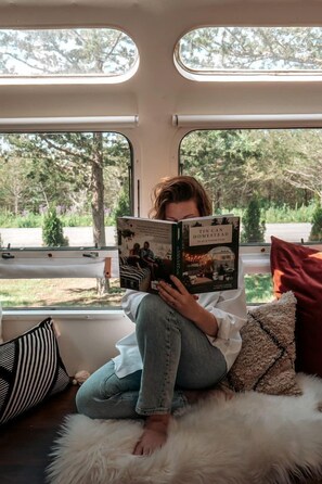 Une personne est assise dans une pièce confortable, lisant un livre. La pièce est dotée d’une grande fenêtre, d’un tapis moelleux et d’un coussin à rayures.
