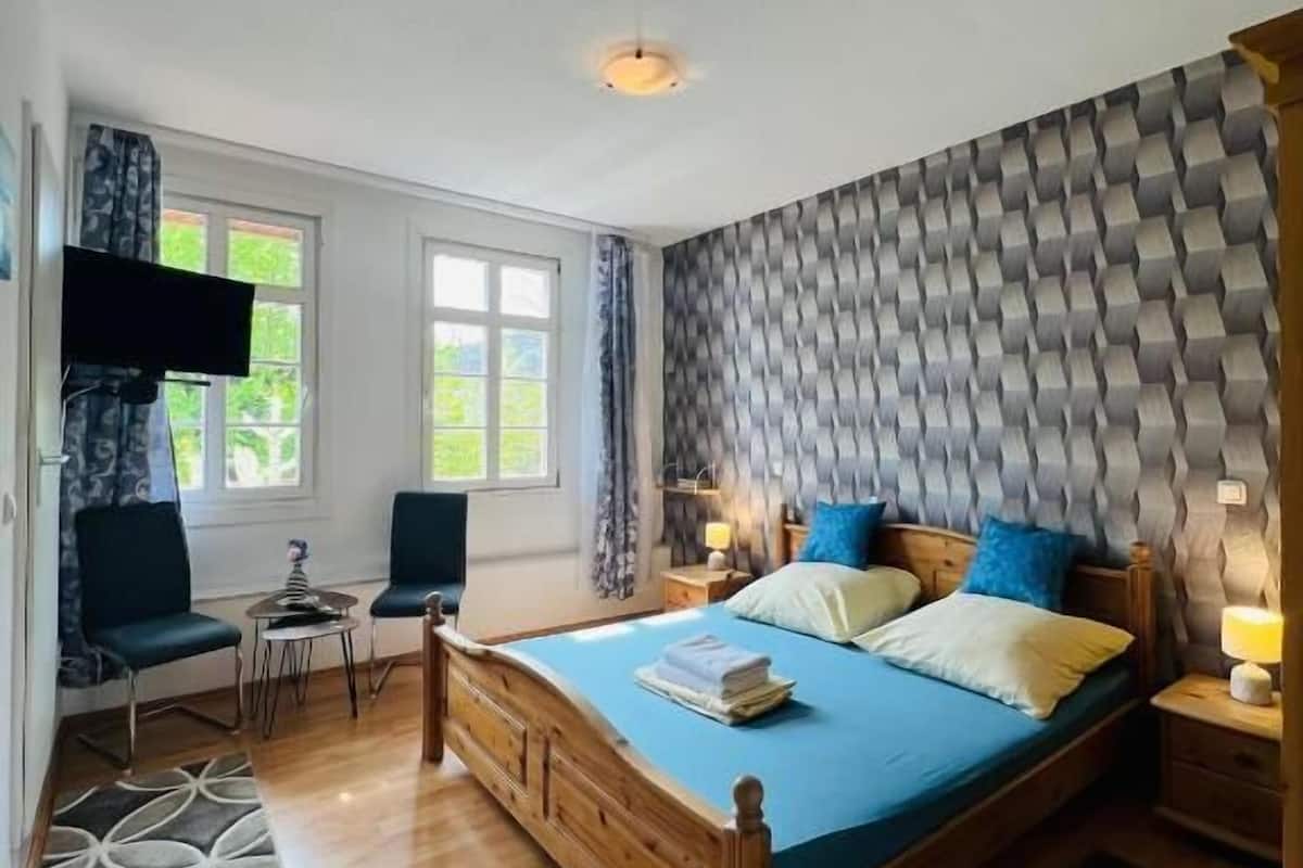 A bedroom with a wooden bed, two bedside tables, a TV mounted on the wall, and a window with curtains.