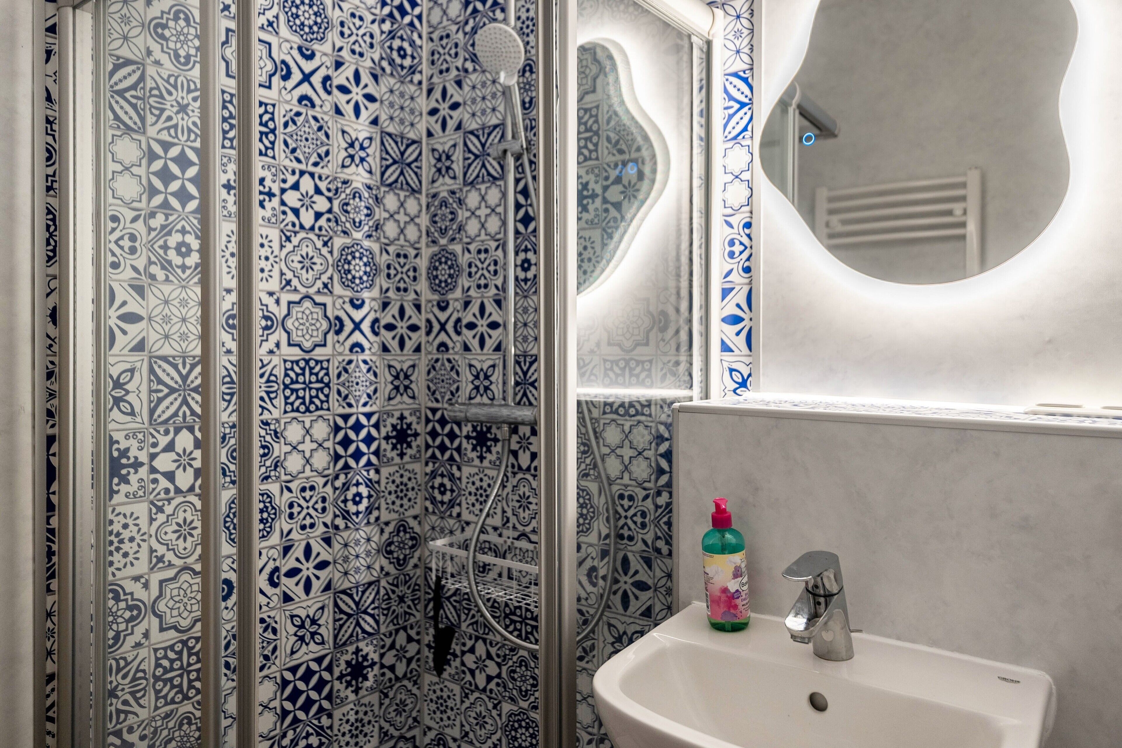 Een badkamer met betegelde vloer, een douche, een wastafel met spiegel en een fles handzeep.