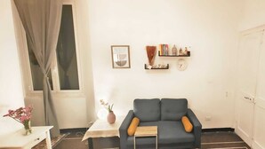 Una sala de estar con un sofá azul oscuro, una mesita con un jarrón de flores, un reloj y una estantería con libros y figuritas.