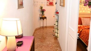 Un pasillo estrecho con una cama a la derecha, una pequeña mesa con una lámpara y un paño rosa, y un espejo a la izquierda.