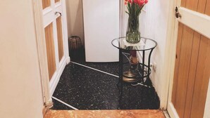 Un pasillo estrecho con piso de mármol negro, un refrigerador blanco, una mesita con una lámpara y un jarrón de flores rojas.