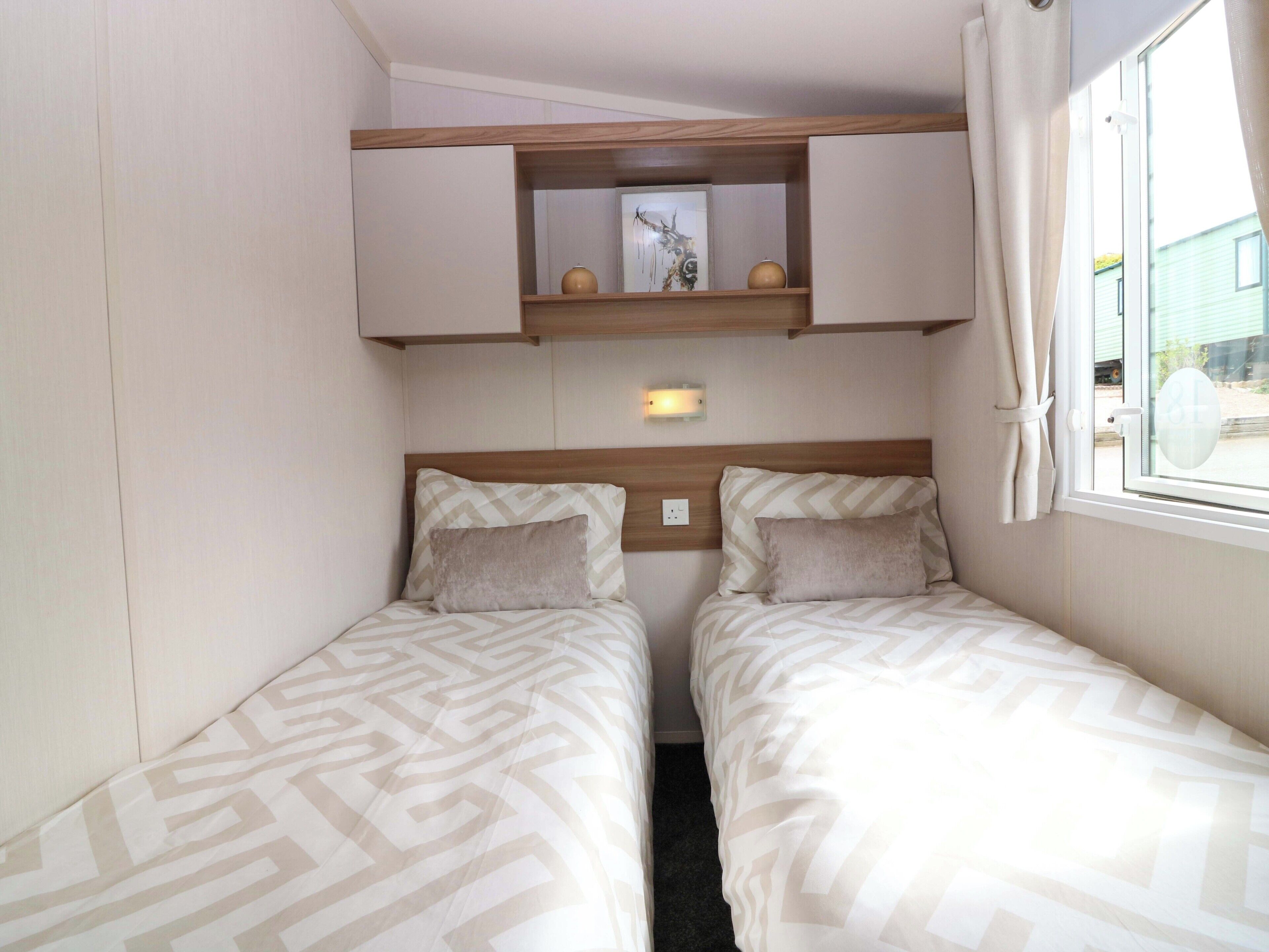 A small room with two beds, a mirror, a shelf with decorative items, and a window with curtains.