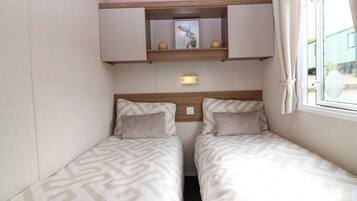 A small room with two beds, a mirror, a shelf with decorative items, and a window with curtains.