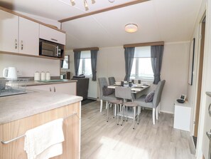 A modern kitchen with a microwave, kettle, and kettle storage. There is a dining area with a table set for four, chairs, and a view of the outdoors through large windows. - 18 Eden Heights (St. Andrews)