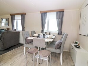 A modern, well-lit room with a dining table set for four, grey upholstered chairs, a grey sofa, and a white cabinet. - 18 Eden Heights (St. Andrews)