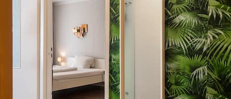 A bedroom with a bed, a nightstand, a wall-mounted light, and a door leading to a room with a tropical wallpaper.